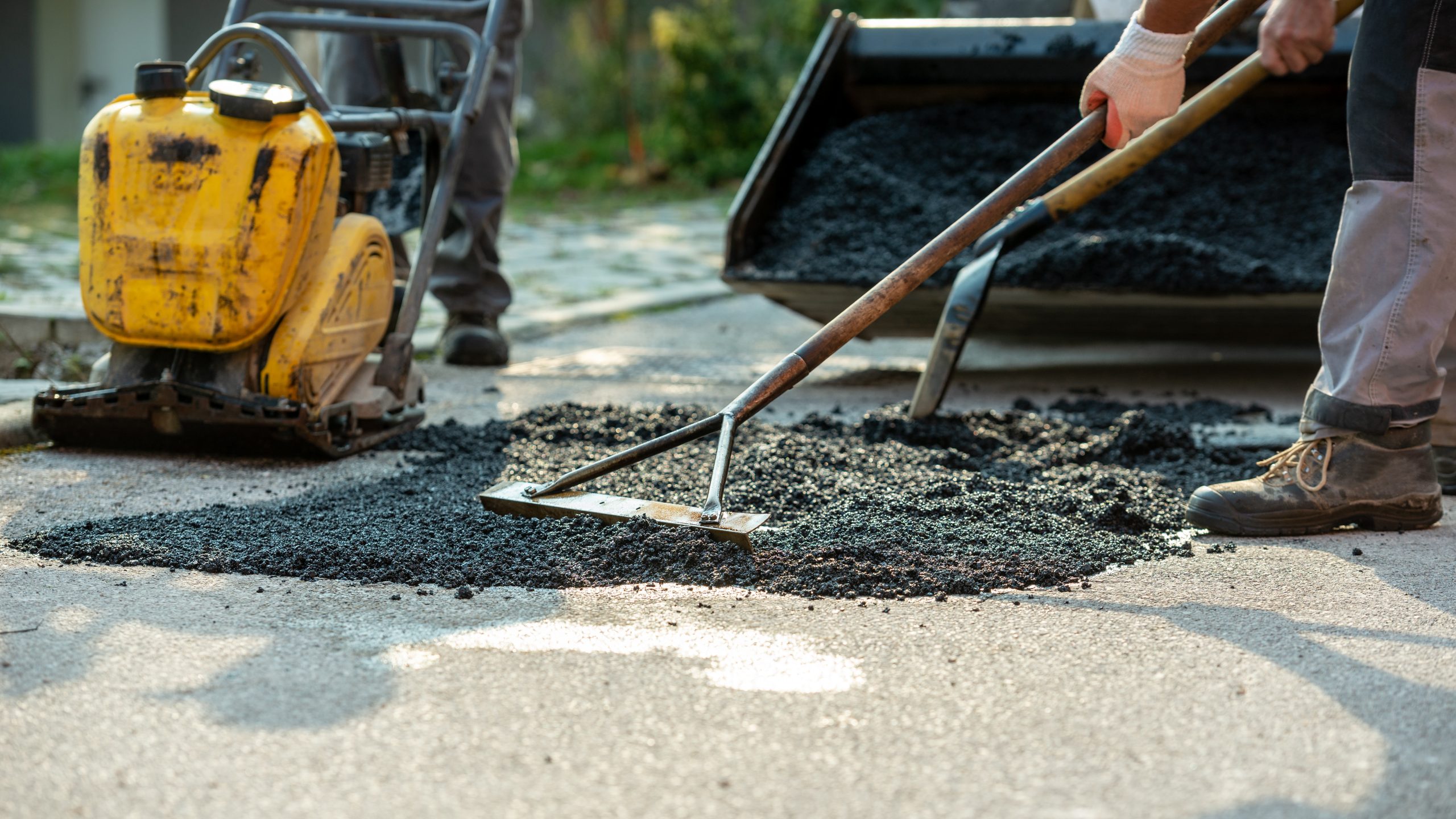 Low angle view of two workers arranging fresh asphalt mix with rakes and shovel to patch a bump in the road with machinery by their side